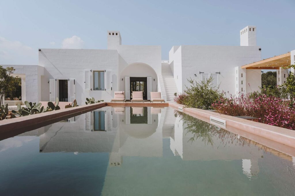 Modern white villa with a reflective pool, surrounded by desert landscaping and vibrant plants, showcasing a serene outdoor living space. | Only on SopranoVillas