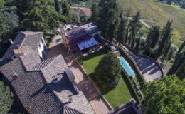 Aerial view of a luxurious villa surrounded by lush greenery, featuring a swimming pool, well-maintained garden, and outdoor seating area, set against a scenic landscape.