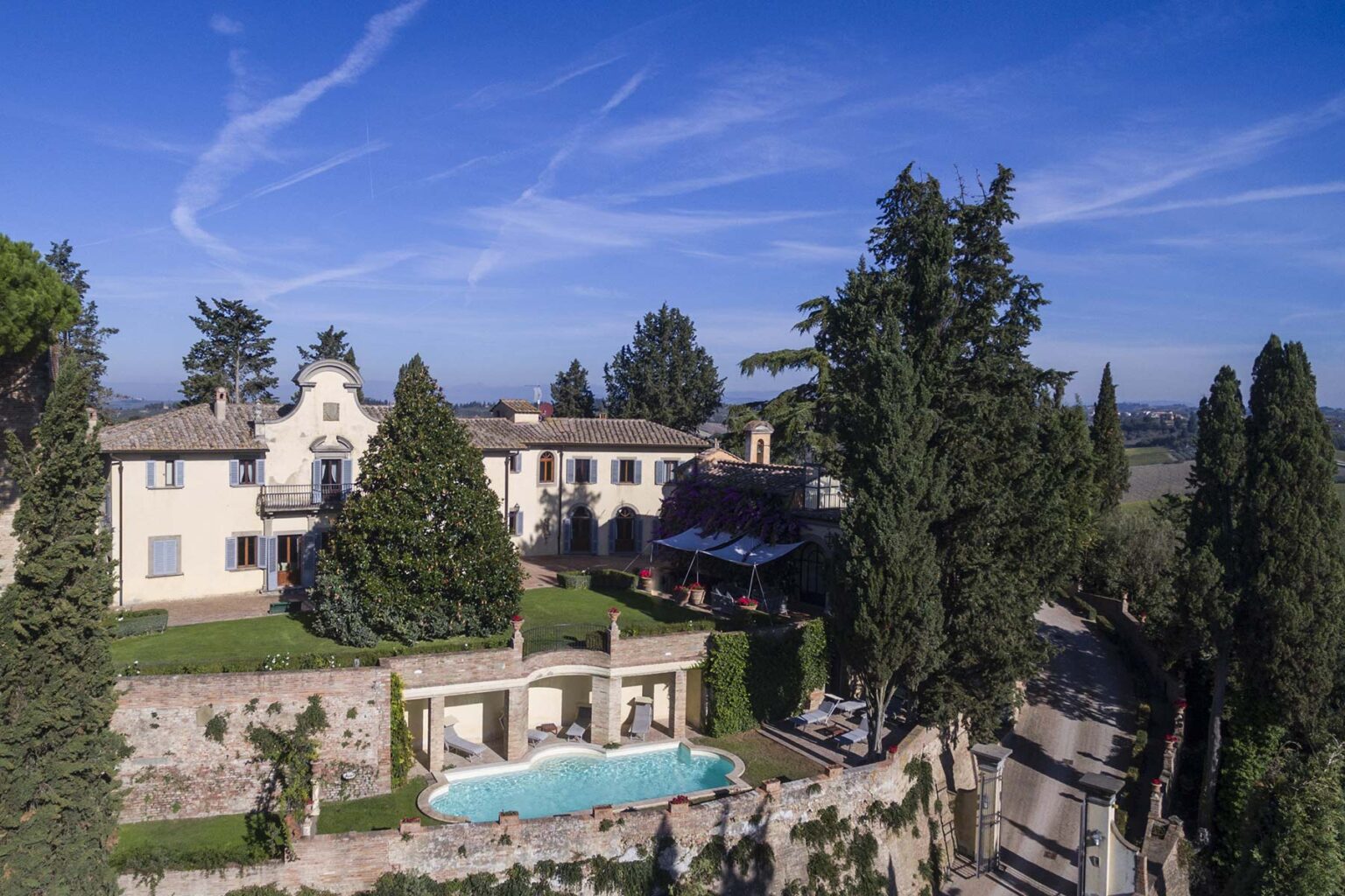 Aerial view of a luxurious villa surrounded by lush greenery, featuring a sparkling swimming pool and elegant architecture against a clear blue sky. | Only on SopranoVillas