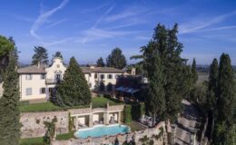 Aerial view of a luxurious villa surrounded by lush greenery, featuring a sparkling swimming pool and elegant architecture against a clear blue sky.