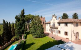 Aerial view of a charming villa surrounded by lush greenery, featuring a swimming pool and well-maintained garden, set against a clear blue sky.