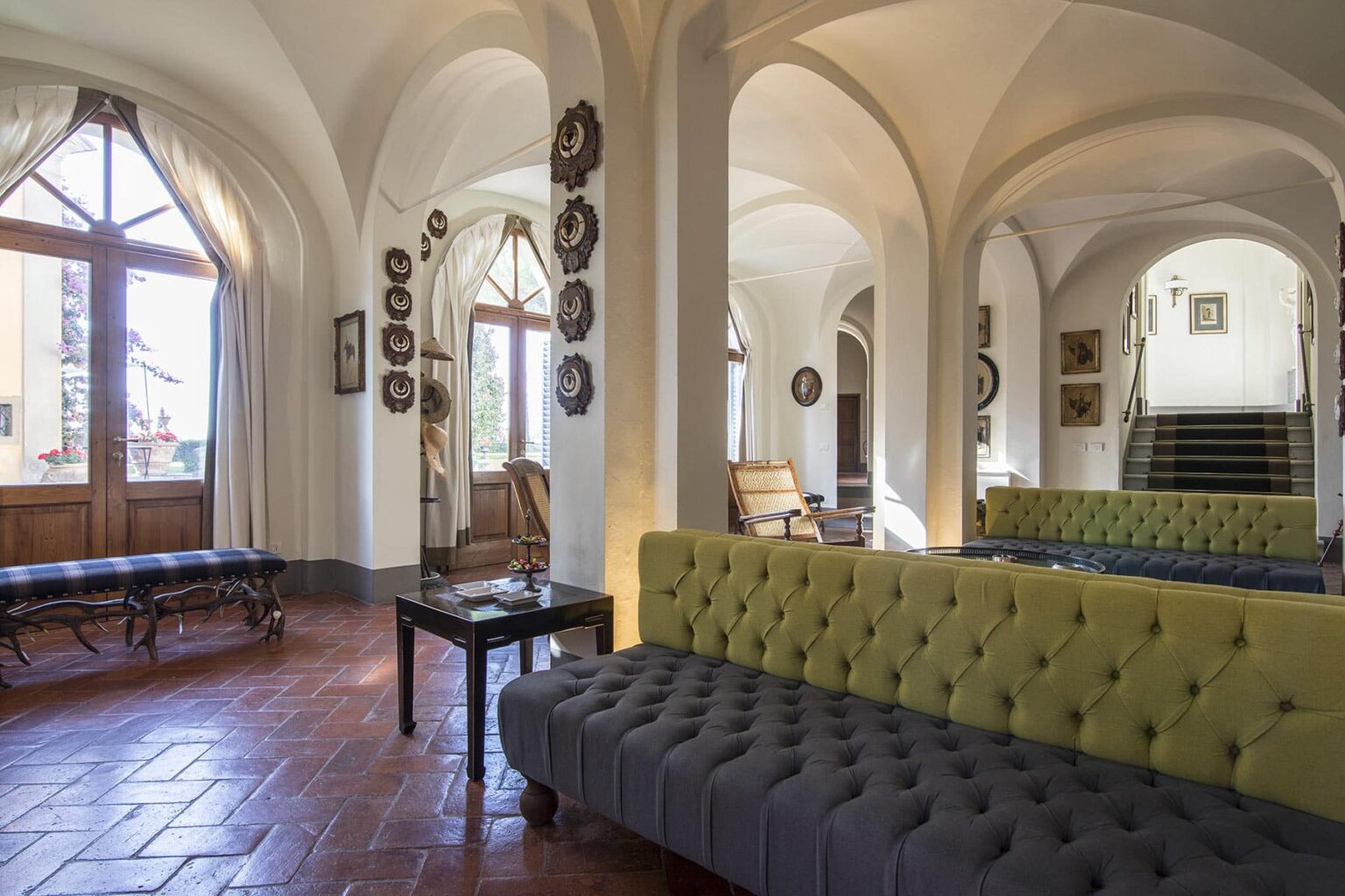 Elegant interior of a spacious lounge featuring arched ceilings, a mix of traditional and modern furniture, and large windows that provide natural light. A gray tufted sofa and a yellow accent sofa are central to the design, complemented by wooden accents and decorative wall art. | Only on SopranoVillas