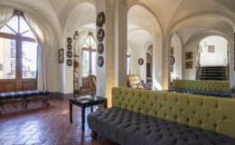 Elegant interior of a spacious lounge featuring arched ceilings, a mix of traditional and modern furniture, and large windows that provide natural light. A gray tufted sofa and a yellow accent sofa are central to the design, complemented by wooden accents and decorative wall art.