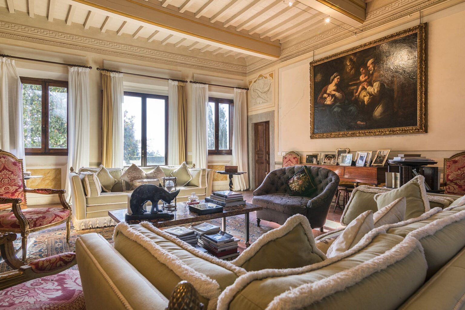 Elegant living room featuring plush sofas, a wooden coffee table, and large windows with natural light. The space is adorned with classic decor, including a large painting and decorative accents, creating a warm and inviting atmosphere. | Only on SopranoVillas