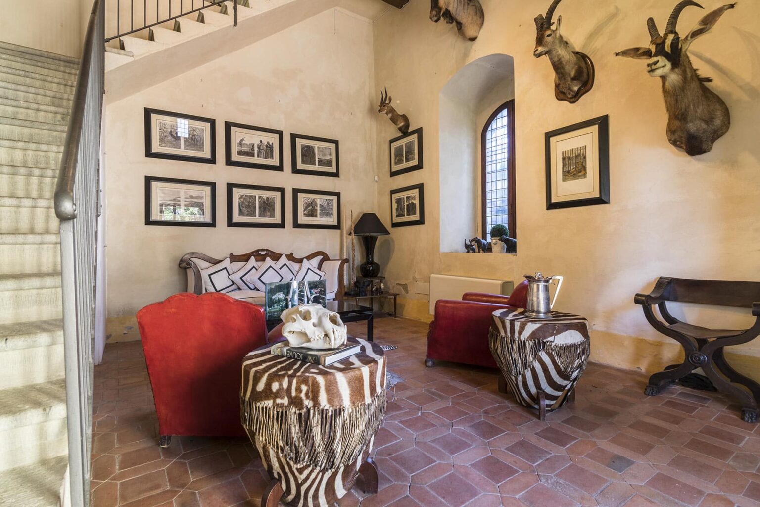 Cozy living room featuring rustic decor, including framed black-and-white photographs on the walls, a comfortable sofa with patterned cushions, and unique zebra-print tables. The space is illuminated by natural light from a large window and has a staircase leading to the upper floor. | Only on SopranoVillas