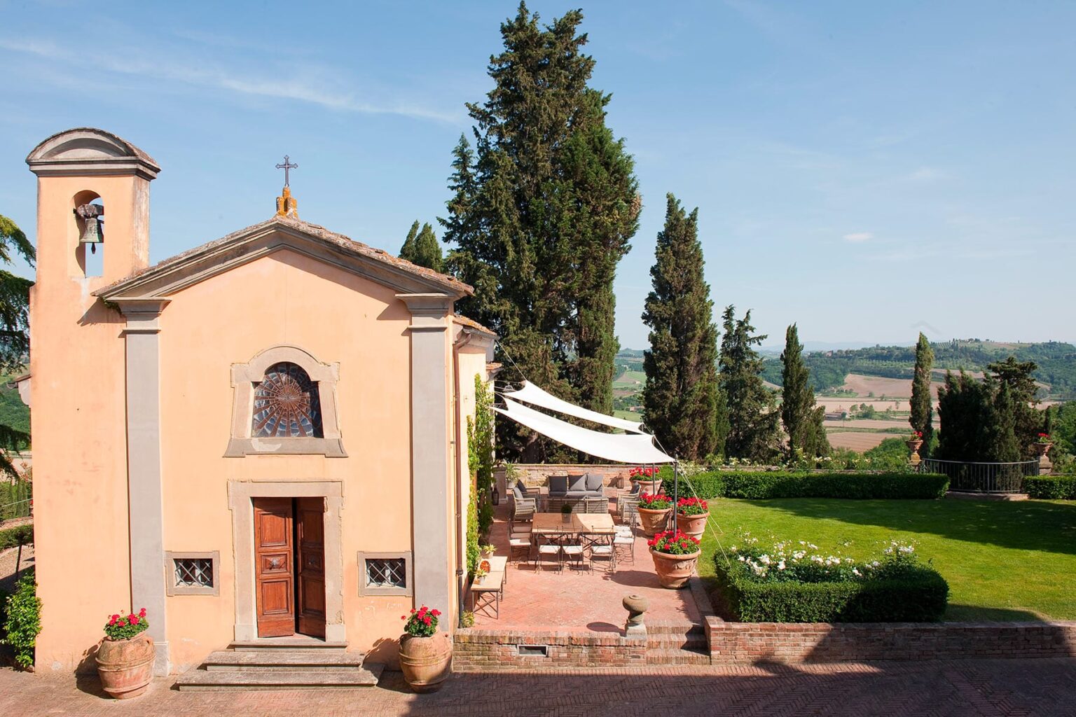 Charming rustic chapel surrounded by lush greenery and cypress trees, featuring a terrace with outdoor seating and beautiful scenic views in the Italian countryside. | Only on SopranoVillas