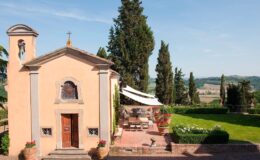 Charming rustic chapel surrounded by lush greenery and cypress trees, featuring a terrace with outdoor seating and beautiful scenic views in the Italian countryside.