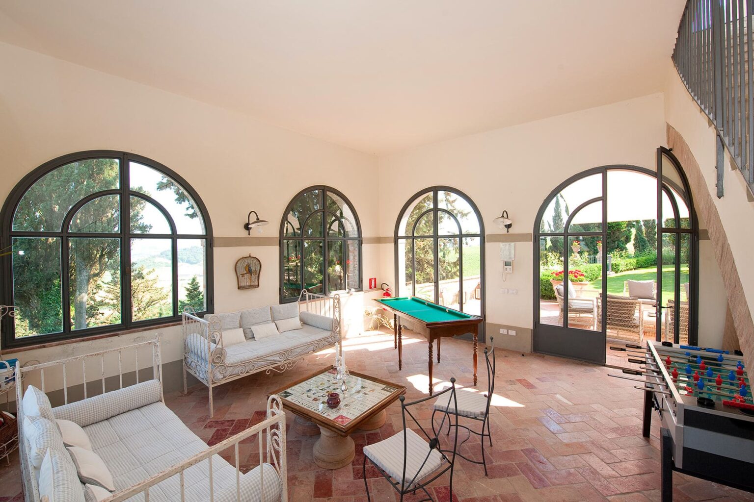 Bright and airy living room featuring large arched windows, a billiard table, and stylish seating arrangements. The space opens to a scenic outdoor view, highlighting a blend of indoor comfort and natural beauty. Perfect for relaxation and entertainment. | Only on SopranoVillas