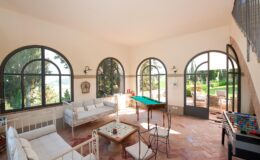 Bright and airy living room featuring large arched windows, a billiard table, and stylish seating arrangements. The space opens to a scenic outdoor view, highlighting a blend of indoor comfort and natural beauty. Perfect for relaxation and entertainment.