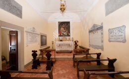 Interior view of a small, historic chapel featuring a central altar, wooden benches, and religious artwork on the walls. The serene atmosphere is enhanced by soft lighting and decorative elements, reflecting traditional architectural style.