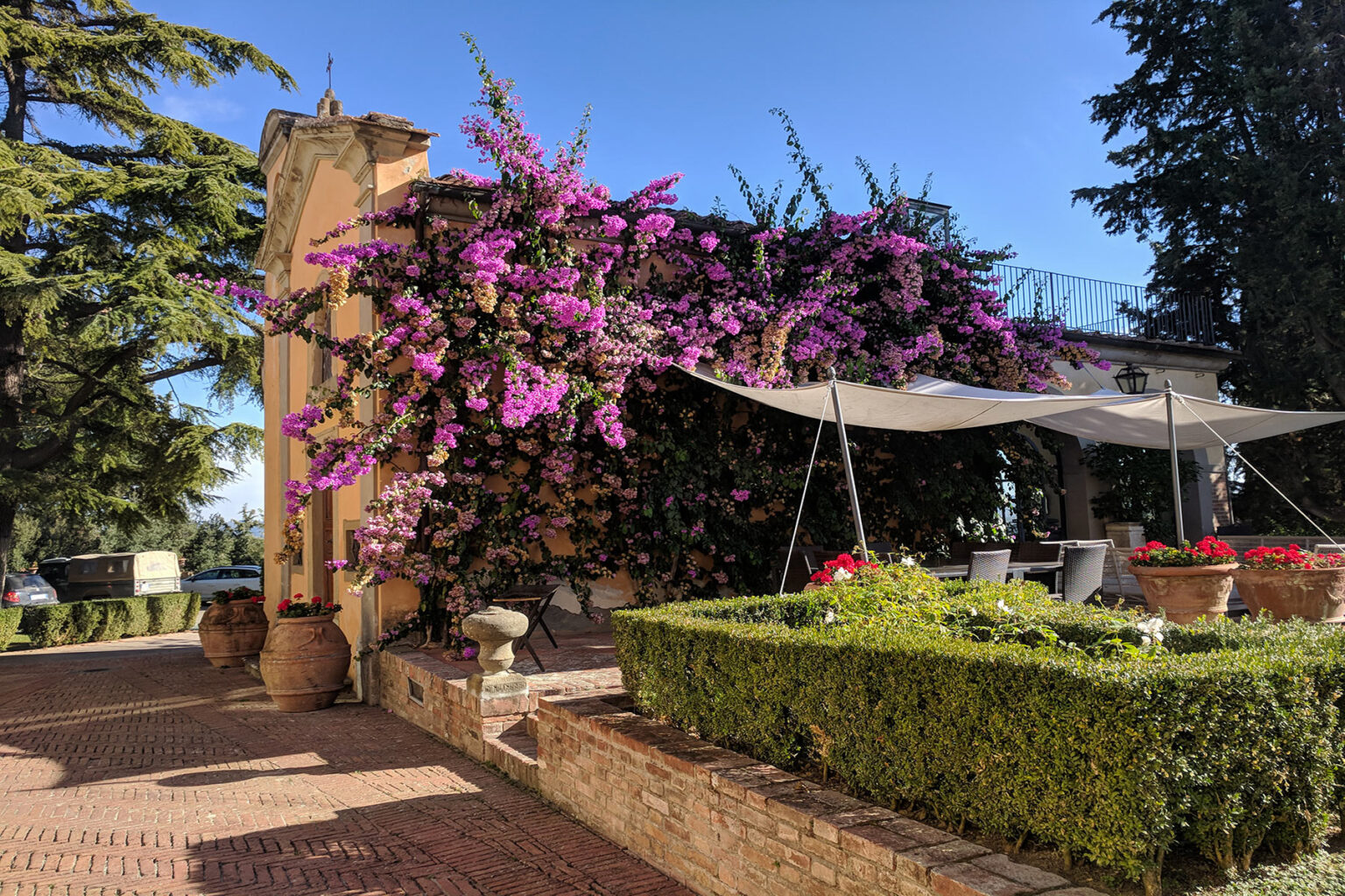Charming outdoor patio with vibrant purple bougainvillea cascading over a stone wall, shaded by a fabric canopy, surrounded by neatly trimmed hedges and colorful flower pots in a sunny garden setting. | Only on SopranoVillas