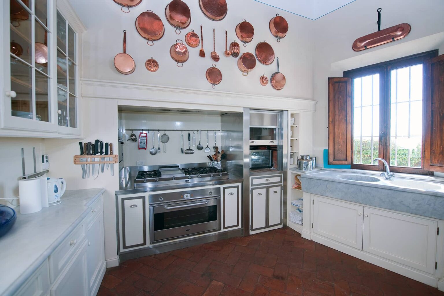 Modern kitchen featuring stainless steel appliances, a marble countertop, and hanging copper pots. Bright natural light streams through large windows, illuminating the spacious layout and cabinetry. | Only on SopranoVillas