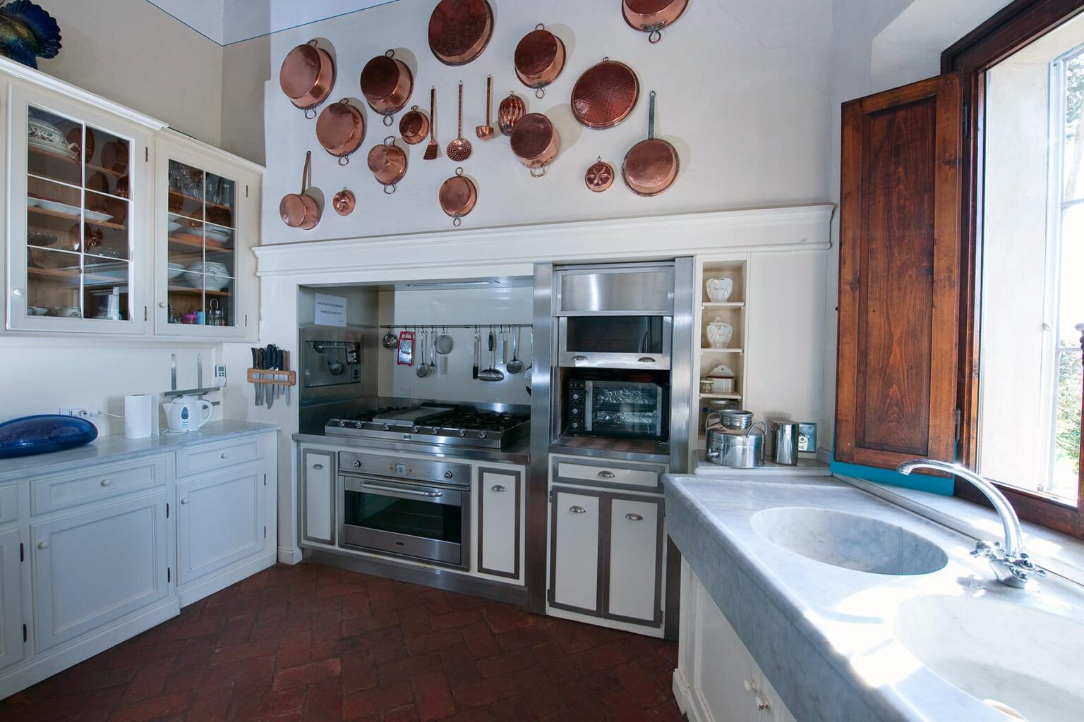 Modern kitchen interior featuring stainless steel appliances, a stone countertop, and wooden cabinetry. Copper pots hang decoratively on the wall, enhancing the rustic charm. Natural light streams in through a window, illuminating the space. | Only on SopranoVillas