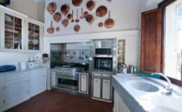 Modern kitchen interior featuring stainless steel appliances, a stone countertop, and wooden cabinetry. Copper pots hang decoratively on the wall, enhancing the rustic charm. Natural light streams in through a window, illuminating the space.
