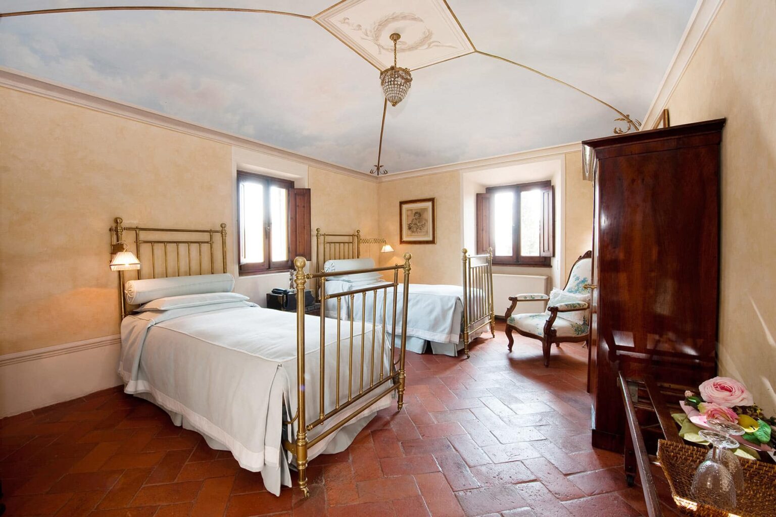 Cozy guest room featuring two brass bed frames, warm terracotta flooring, and elegant decor. Natural light floods the space through large windows, creating a welcoming atmosphere. Ideal for relaxation and comfort during your stay. | Only on SopranoVillas