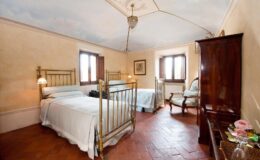 Cozy guest room featuring two brass bed frames, warm terracotta flooring, and elegant decor. Natural light floods the space through large windows, creating a welcoming atmosphere. Ideal for relaxation and comfort during your stay.