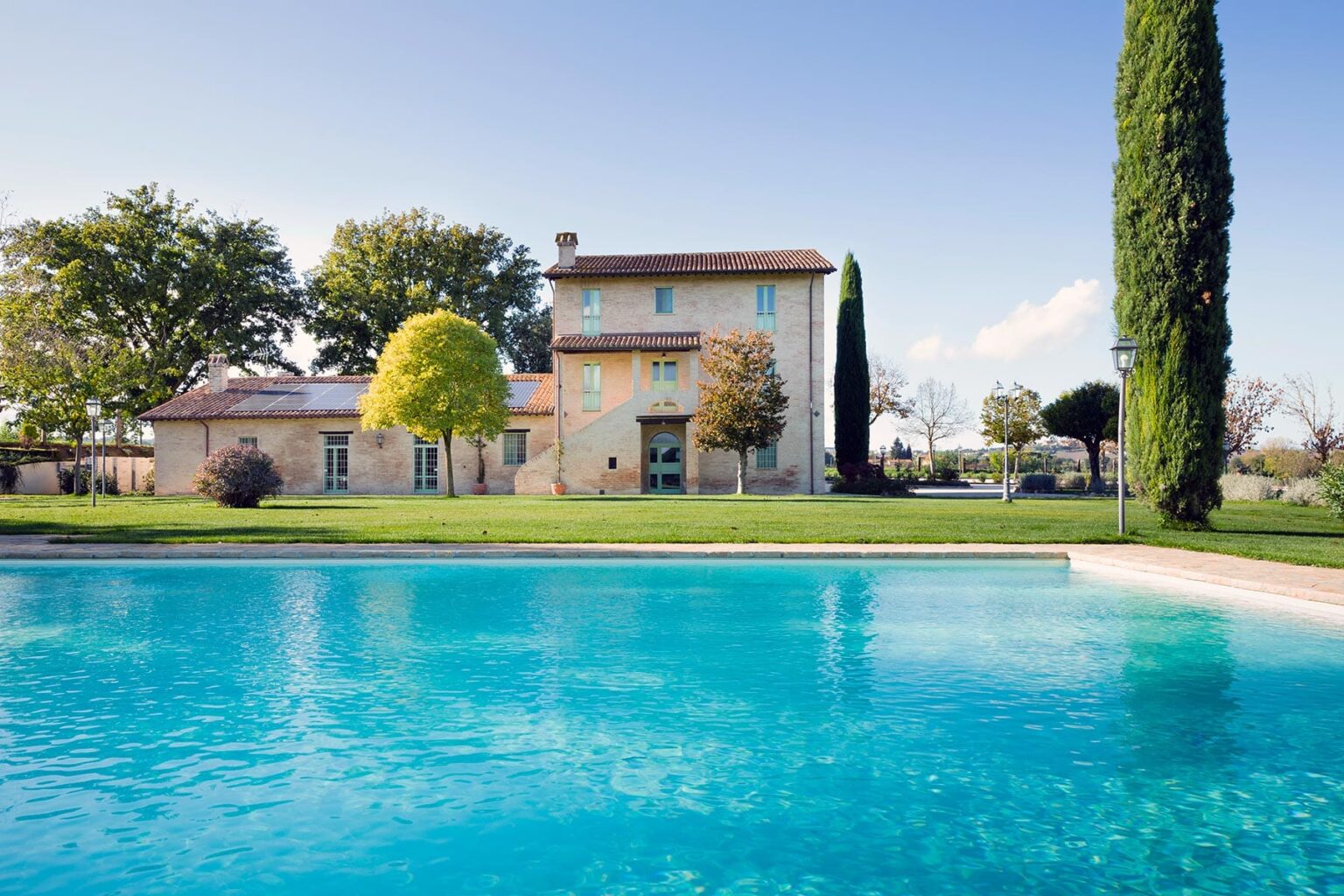 Elegant villa with a swimming pool surrounded by lush greenery and tall cypress trees, set against a clear blue sky. Ideal for luxury retreats and vacation rentals. | Only on SopranoVillas