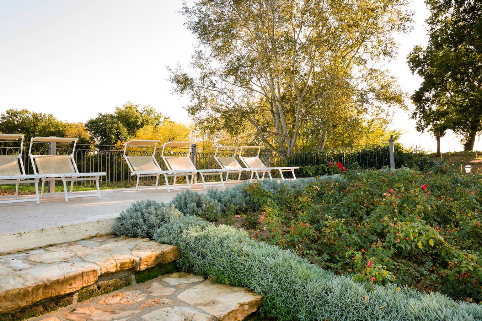 Outdoor seating area with lounge chairs beside a landscaped garden, featuring lush greenery and blooming flowers, illuminated by warm sunlight. | Only on SopranoVillas