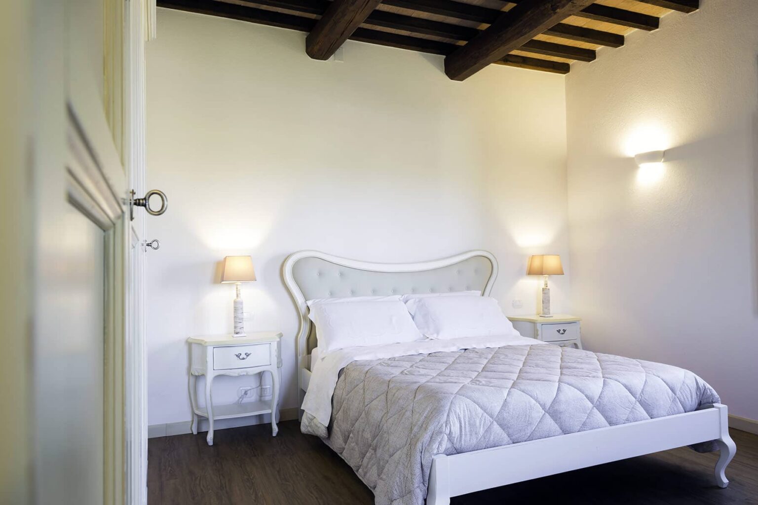 Cozy bedroom featuring a queen-sized bed with a light gray quilt, elegant bedside tables with lamps, and exposed wooden beams on the ceiling, creating a warm and inviting atmosphere. | Only on SopranoVillas