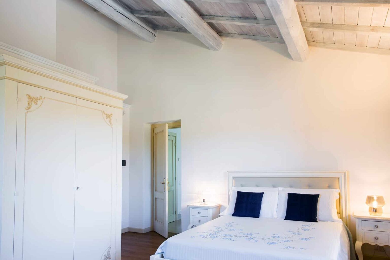 Bright and airy bedroom featuring a cozy bed with white linens and navy blue pillows, complemented by a vintage wardrobe and soft lighting, showcasing a serene and inviting atmosphere. | Only on SopranoVillas