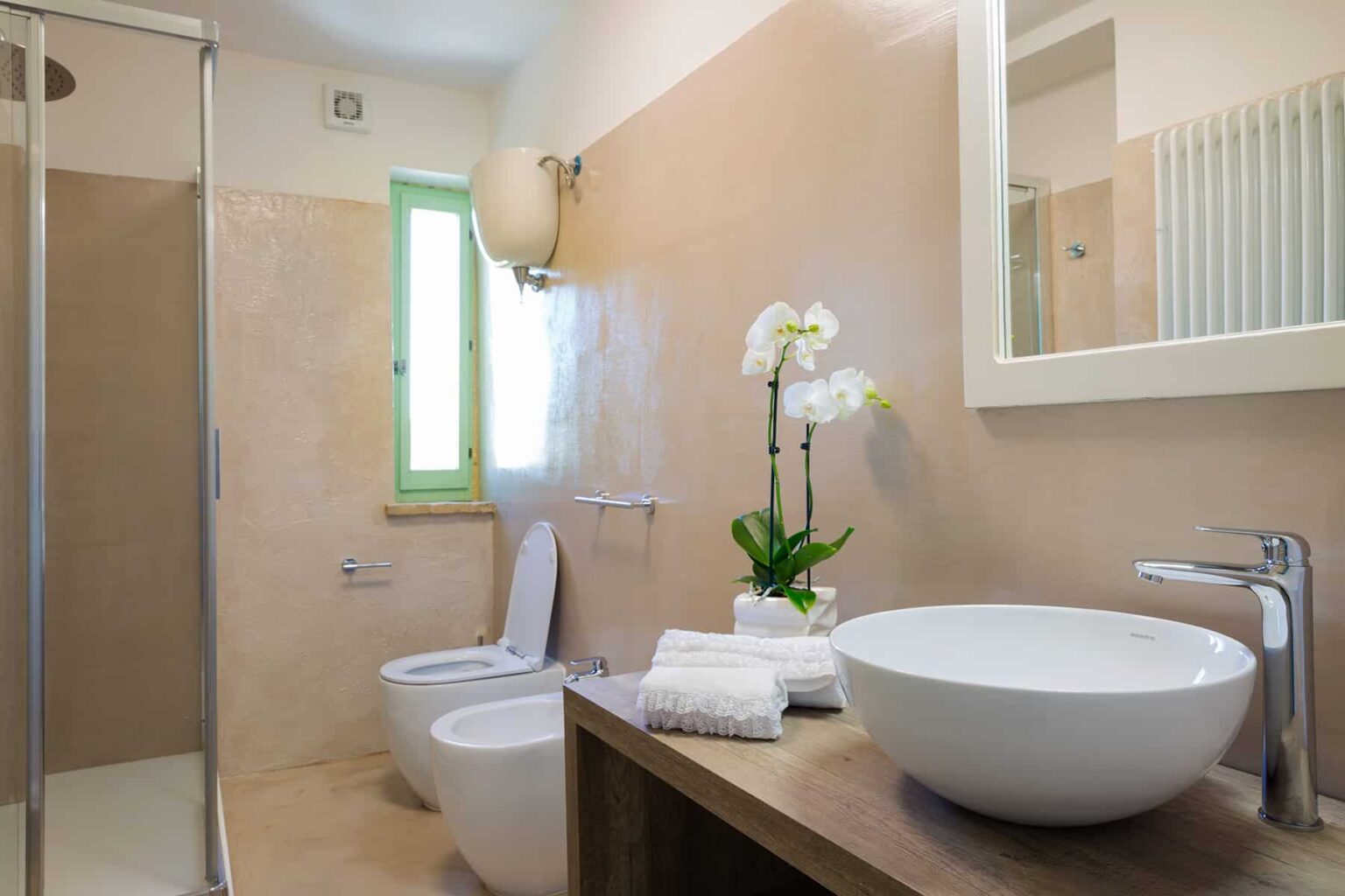 Modern bathroom featuring a sleek design with a glass shower, elegant bowl sink, and natural light from a green-framed window. Decorated with white towels and a potted orchid for a touch of sophistication. | Only on SopranoVillas