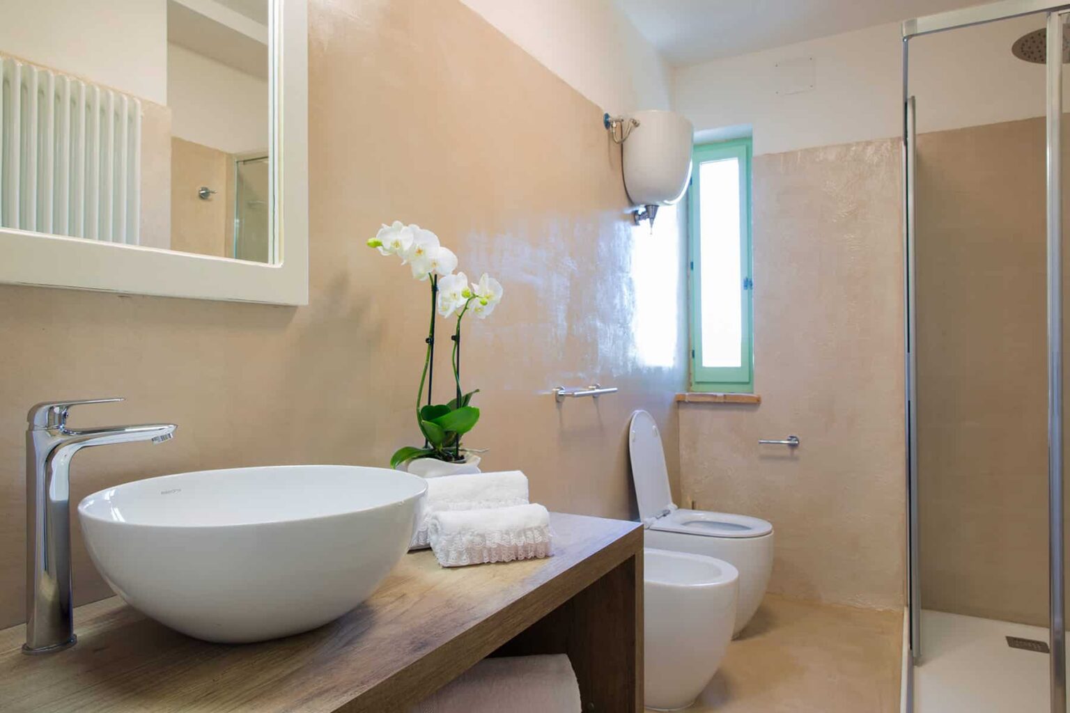 Modern bathroom design featuring a round vessel sink on a wooden countertop, a white toilet, and a glass shower. Natural light enters through a green-framed window, complemented by decorative orchids and neatly arranged towels. | Only on SopranoVillas