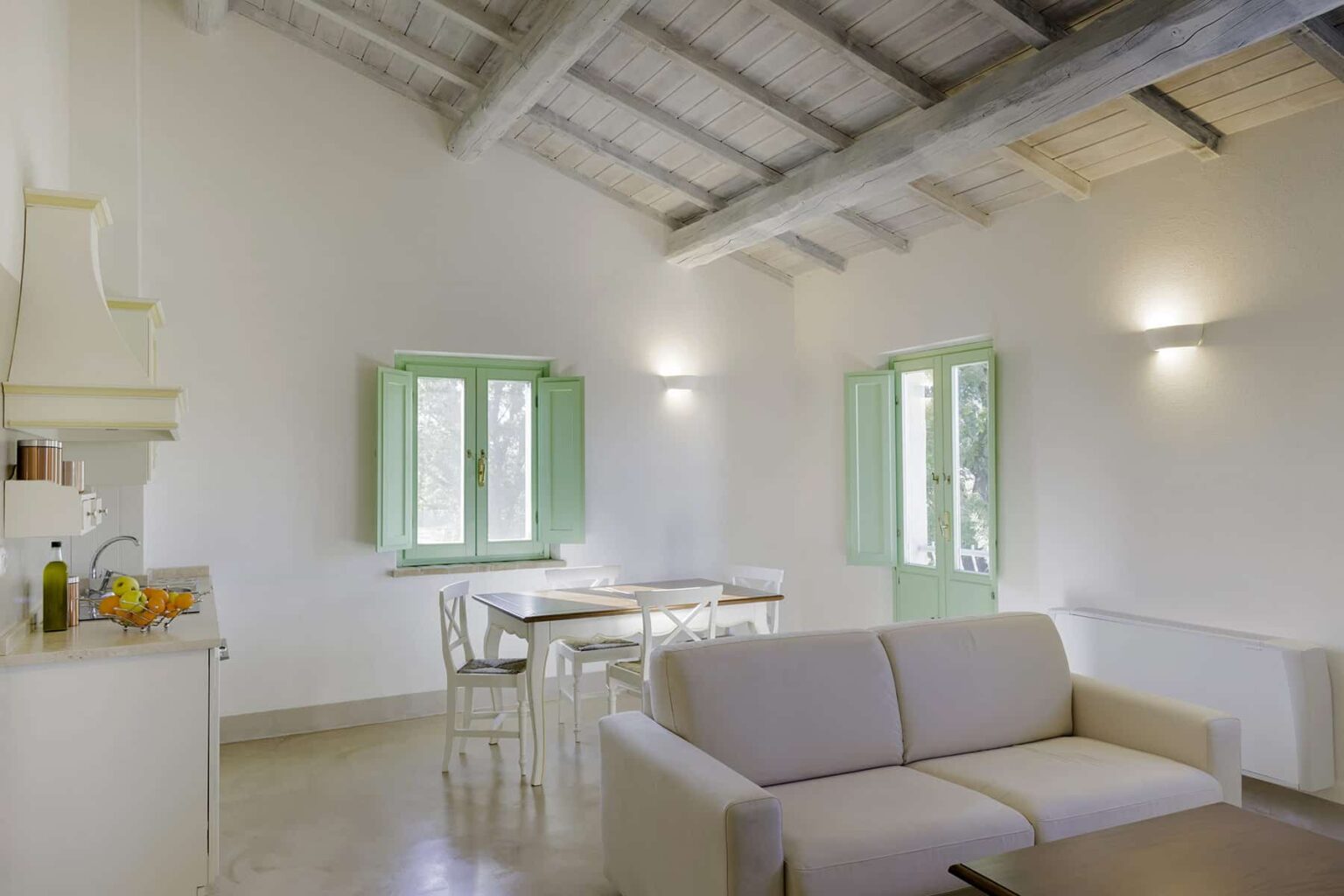 Bright and airy living space featuring a modern design with a white color palette, wooden ceiling beams, a comfortable sofa, and a dining area with green window frames. Ideal for relaxation and entertaining. | Only on SopranoVillas