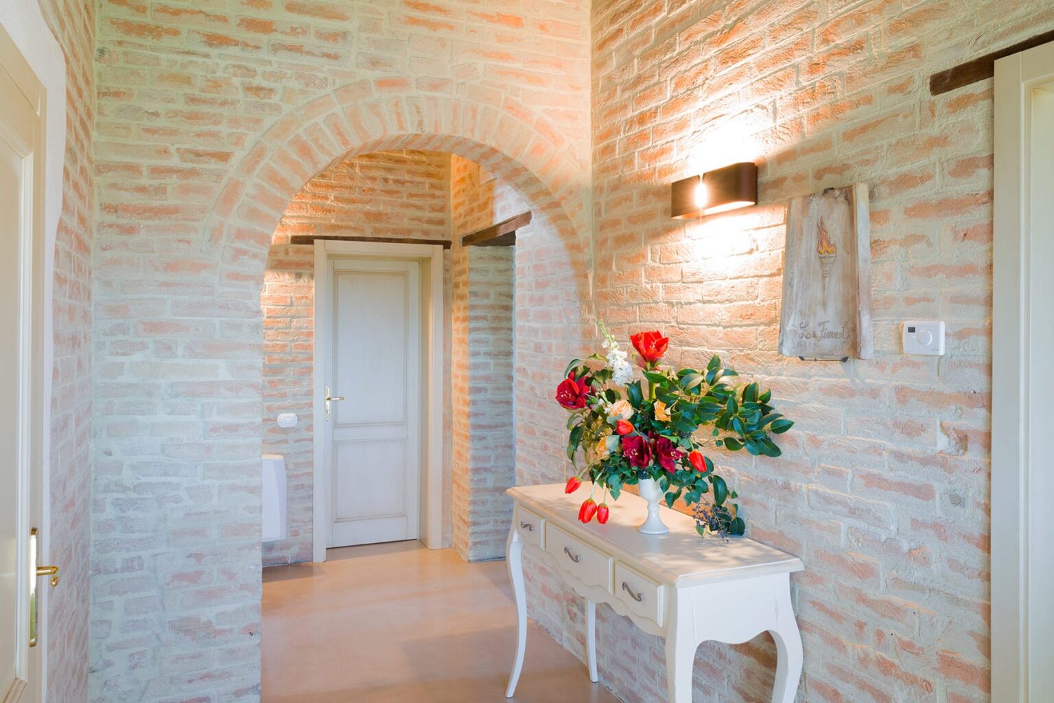 Interior view of a stylish hallway featuring a brick archway, decorative lighting, and a white console table adorned with red roses and greenery, creating an inviting atmosphere. | Only on SopranoVillas