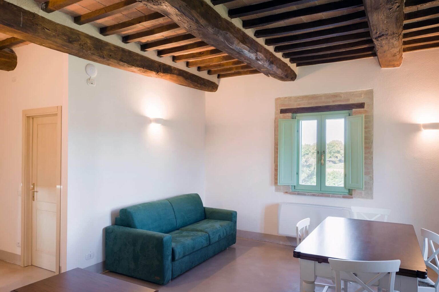 Cozy living room featuring a green sofa, wooden beam ceiling, and a large window with shutters, creating a bright and inviting atmosphere. | Only on SopranoVillas