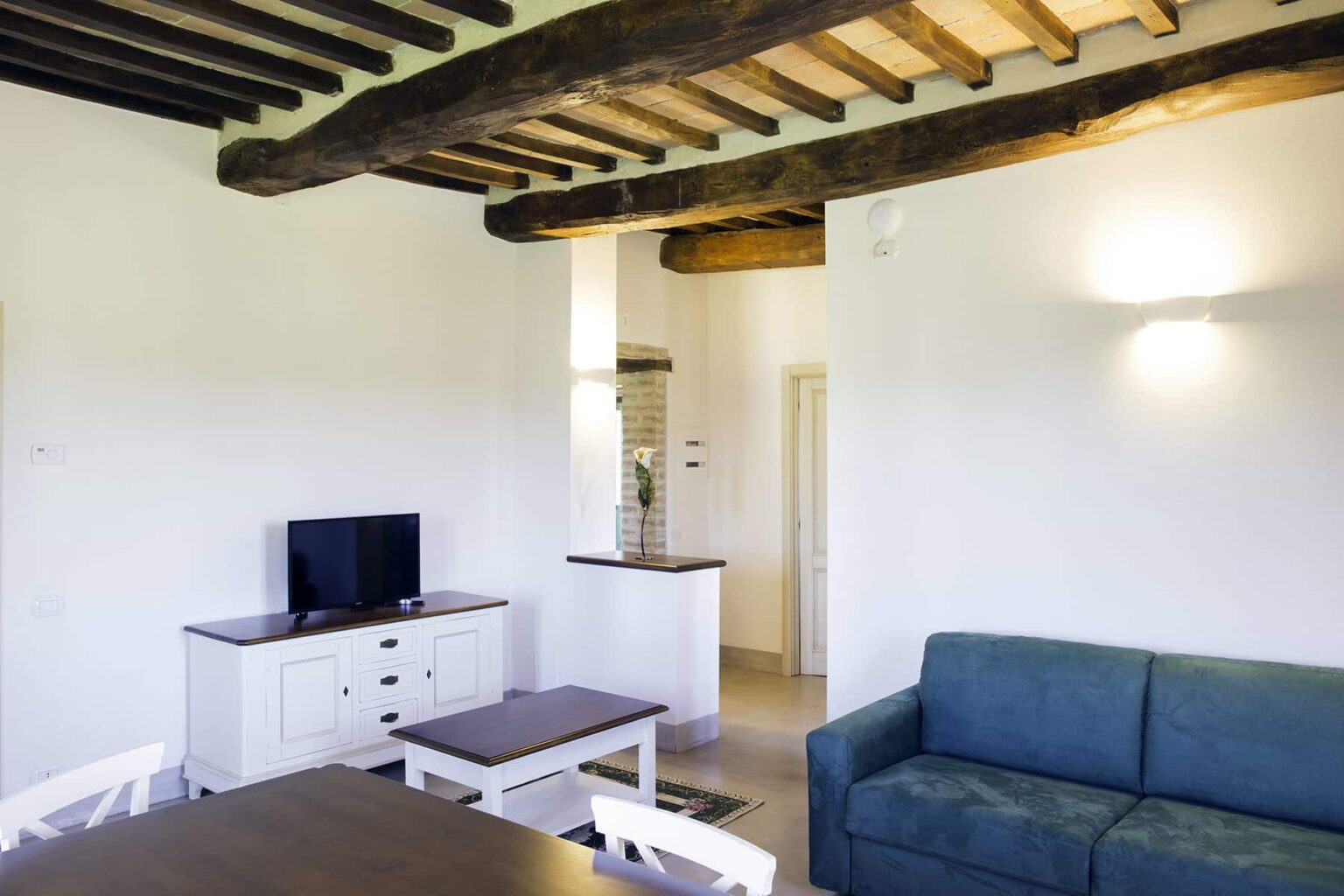Modern living room interior featuring a blue sofa, wooden ceiling beams, a white TV cabinet, and a coffee table, creating a cozy and inviting atmosphere. | Only on SopranoVillas