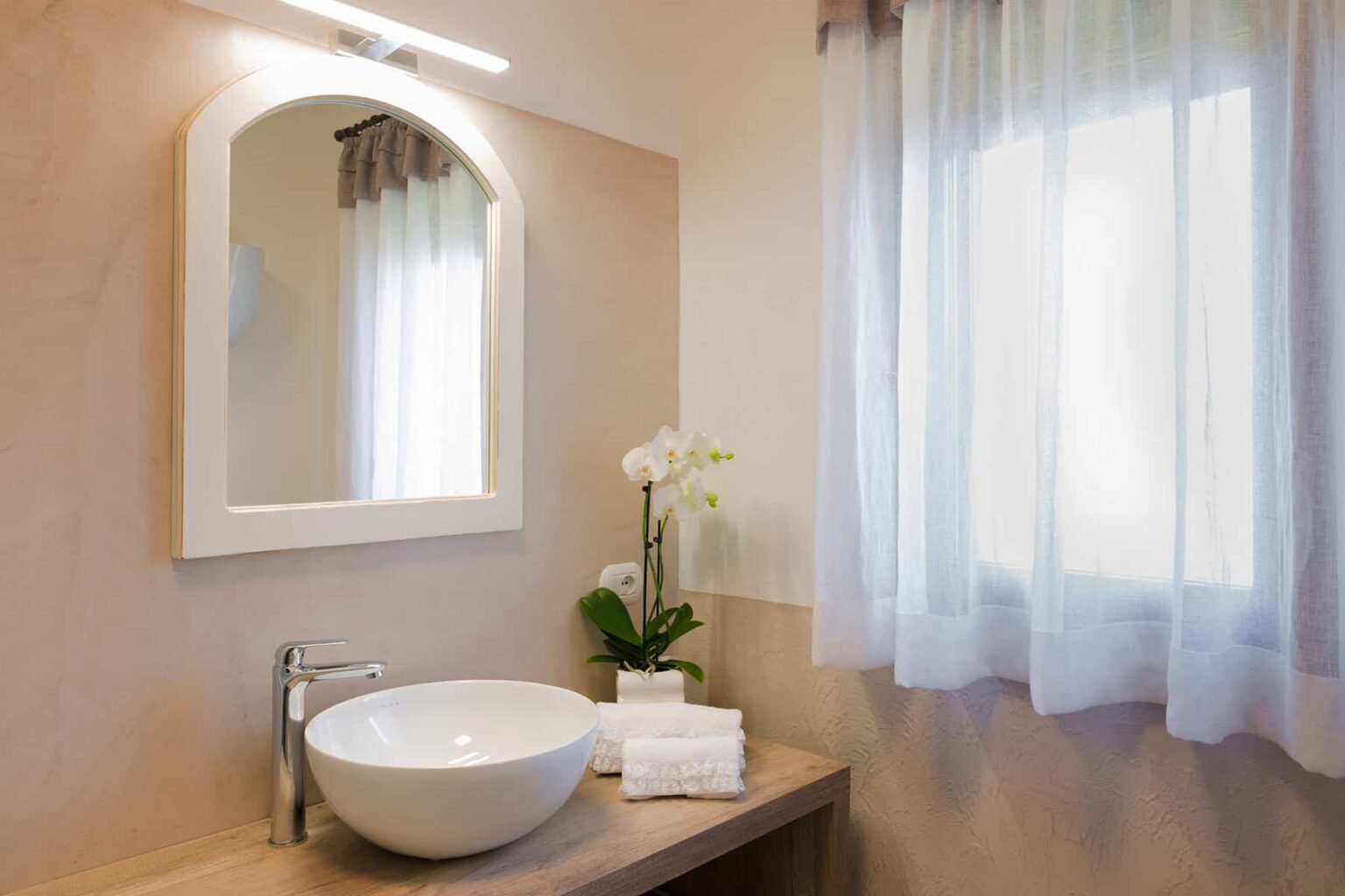Modern bathroom interior featuring a round vessel sink, stylish mirror, and natural light from a window. Decorated with a potted orchid and neatly arranged towels, creating a serene and elegant atmosphere. | Only on SopranoVillas