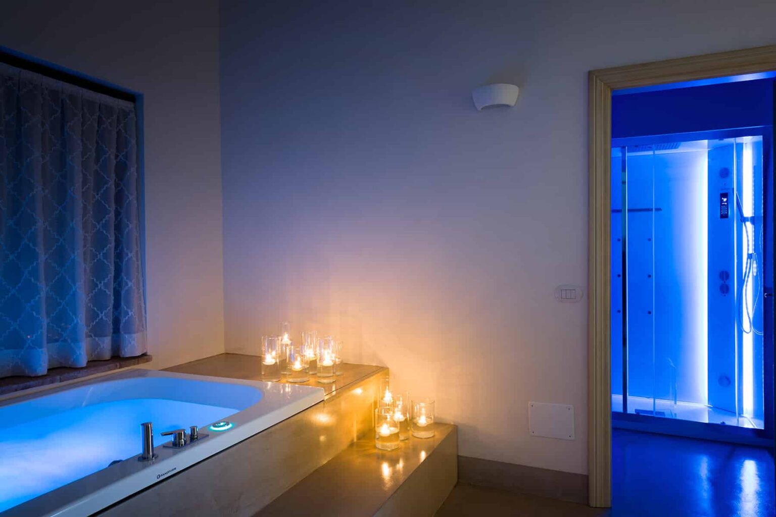 Modern bathroom featuring a relaxing bathtub illuminated by candles, with a soothing blue light from a nearby shower area, creating a tranquil spa-like atmosphere. | Only on SopranoVillas