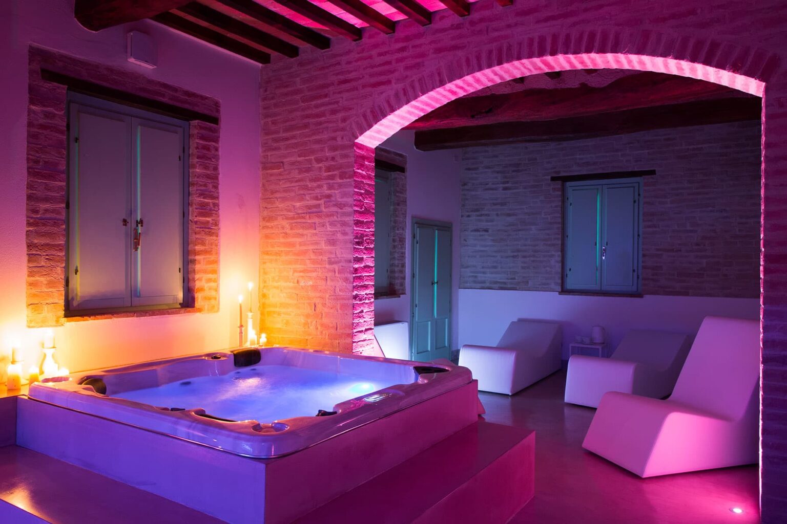 Modern spa room featuring a luxurious hot tub illuminated with colorful LED lights, surrounded by minimalist furniture and exposed brick walls, creating a relaxing ambiance. | Only on SopranoVillas