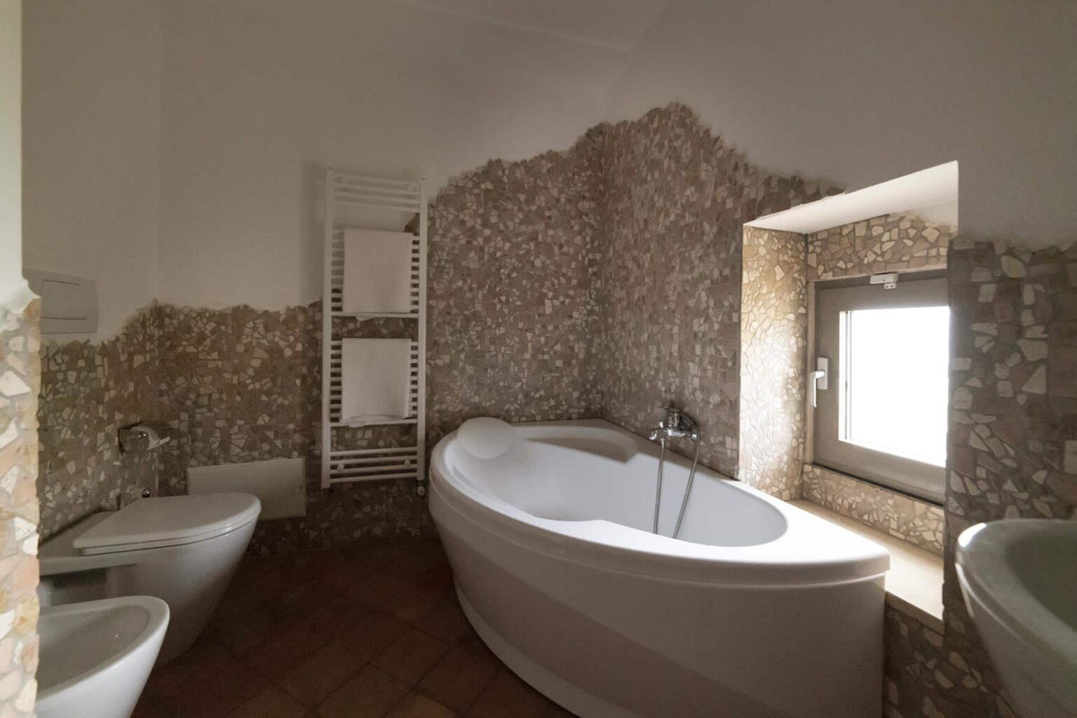 Modern bathroom interior featuring a stylish freestanding bathtub, a bidet, and a window allowing natural light, with textured stone walls and wooden flooring for a contemporary aesthetic. | Only on SopranoVillas