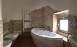 Modern bathroom interior featuring a stylish freestanding bathtub, a bidet, and a window allowing natural light, with textured stone walls and wooden flooring for a contemporary aesthetic.