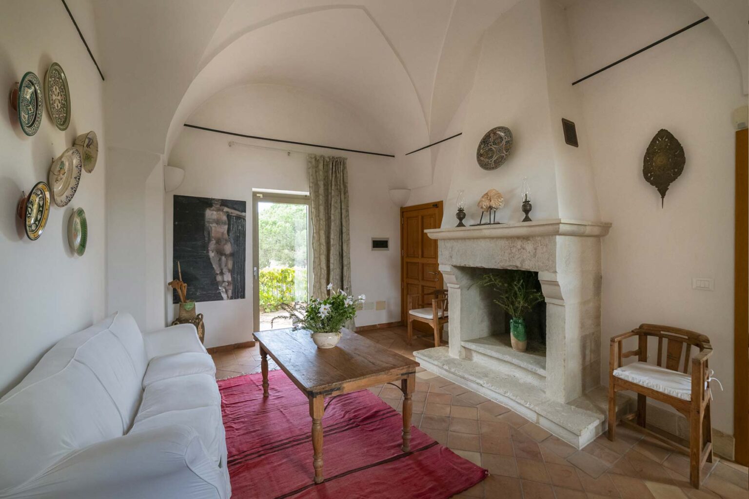 Cozy living room featuring a white sofa, wooden coffee table, and a stone fireplace, adorned with decorative plates and artwork, complemented by natural light from the window. | Only on SopranoVillas
