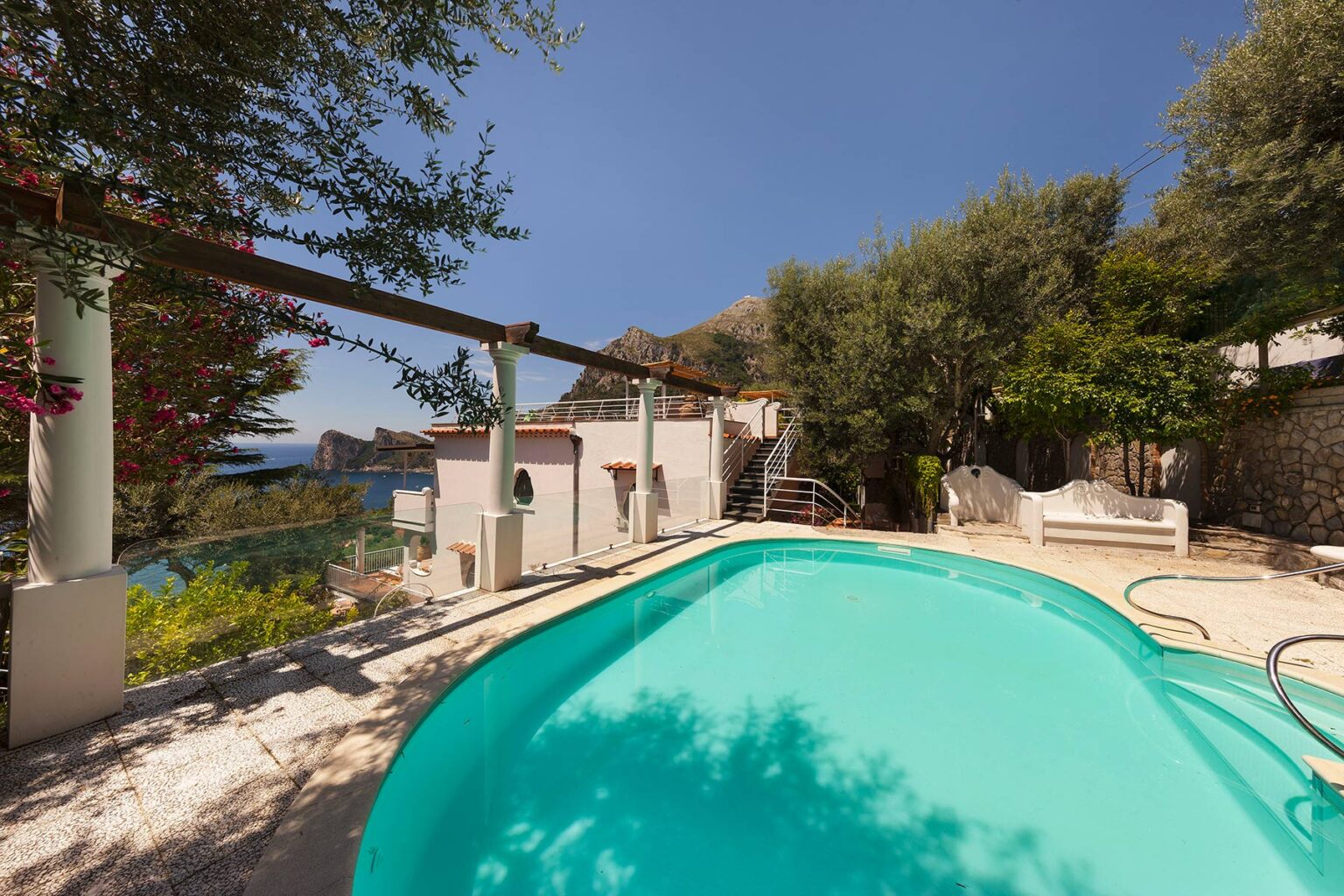 A serene swimming pool surrounded by lush greenery and a modern villa, set against a clear blue sky. | Only on SopranoVillas
