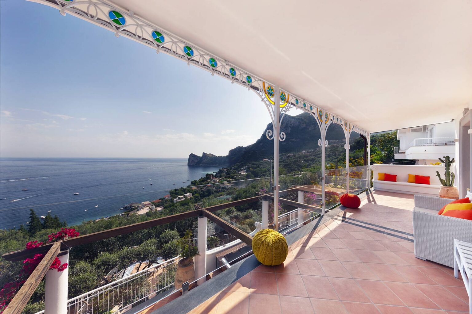 Stunning coastal view from a modern terrace overlooking a serene ocean and mountainous landscape, featuring stylish outdoor seating and vibrant decor elements. | Only on SopranoVillas