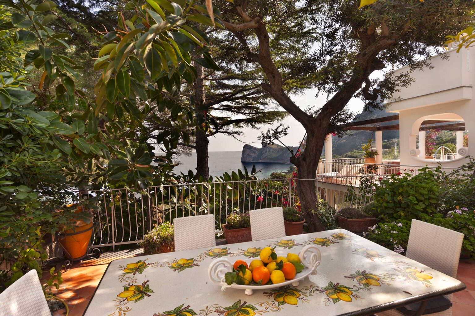 Outdoor dining area featuring a table adorned with fresh fruits, surrounded by lush greenery and trees, with a view of a nearby building. Ideal for relaxing meals in a serene garden setting. | Only on SopranoVillas