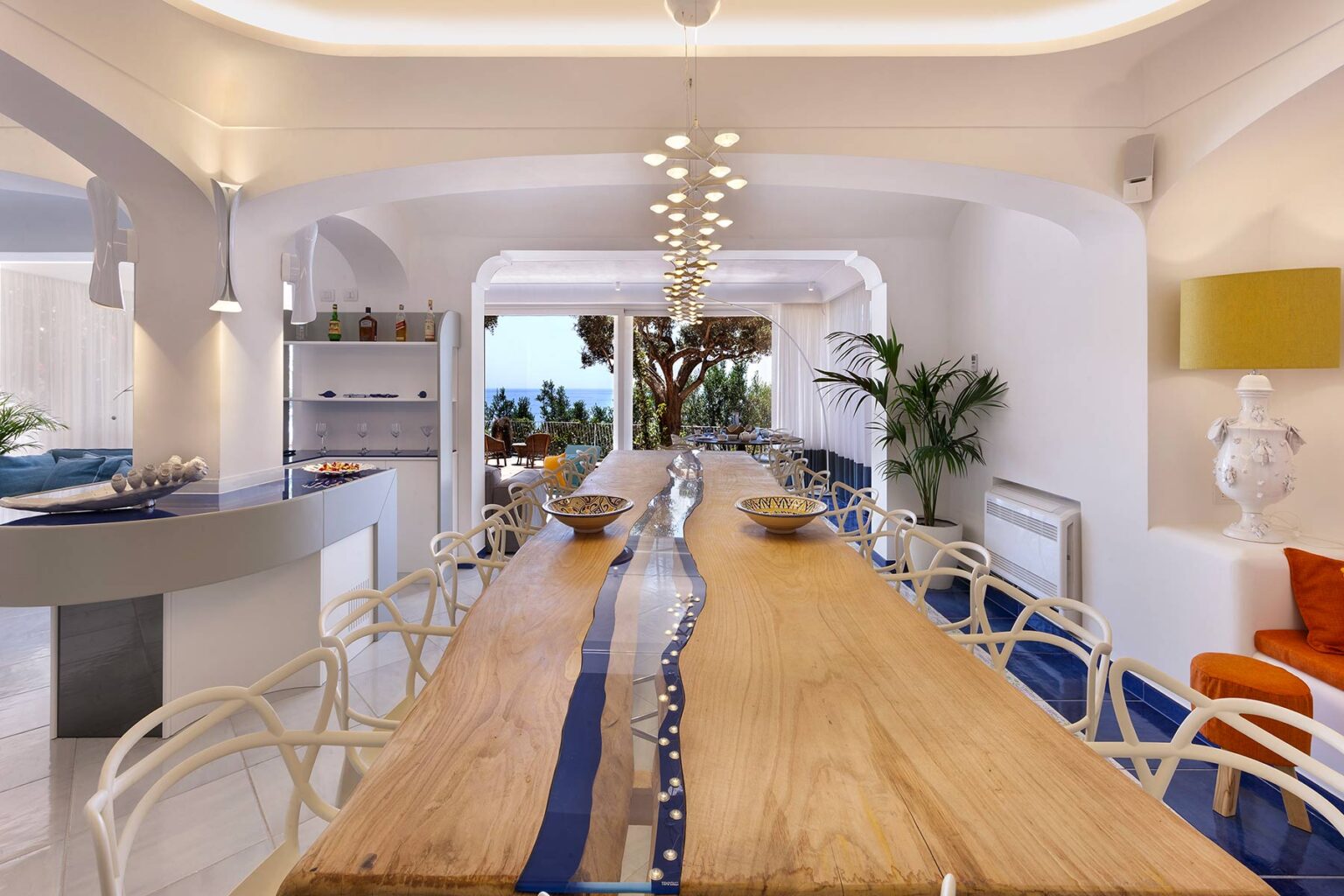 Modern dining area featuring a long wooden table with a blue river design, surrounded by stylish white chairs. The space is illuminated with contemporary pendant lights and decorated with plants, offering a bright and inviting atmosphere. | Only on SopranoVillas