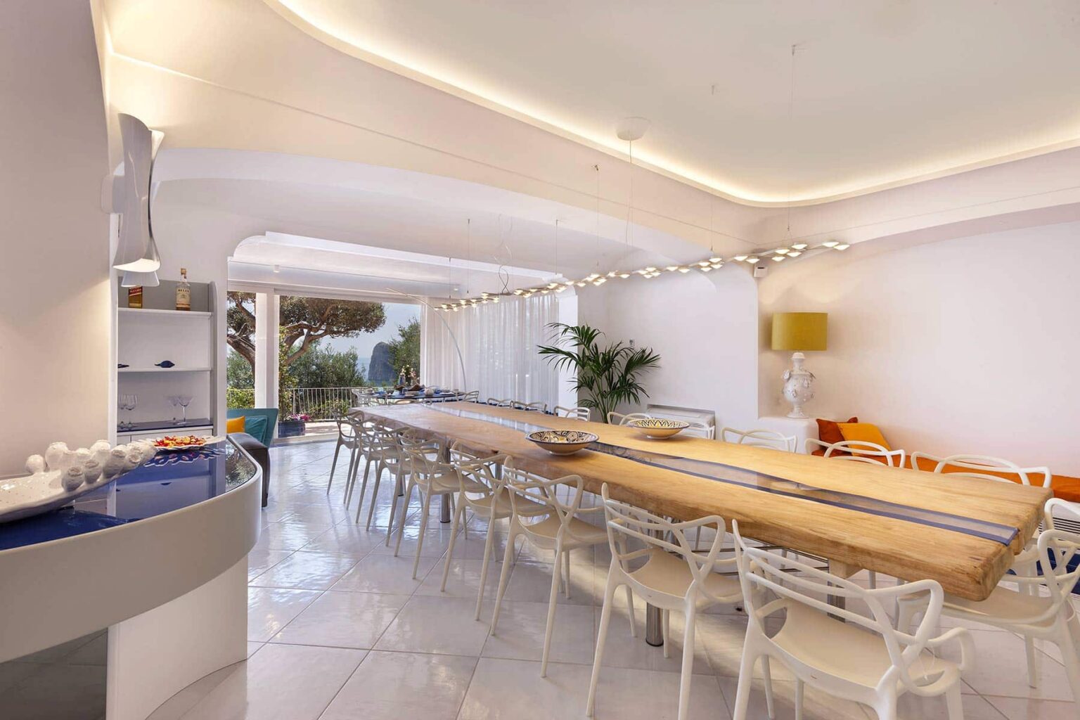 Spacious modern dining area featuring a long wooden table, transparent chairs, and large windows that provide natural light and a view of greenery. The sleek design includes white walls and contemporary lighting fixtures, creating an inviting atmosphere for gatherings. | Only on SopranoVillas