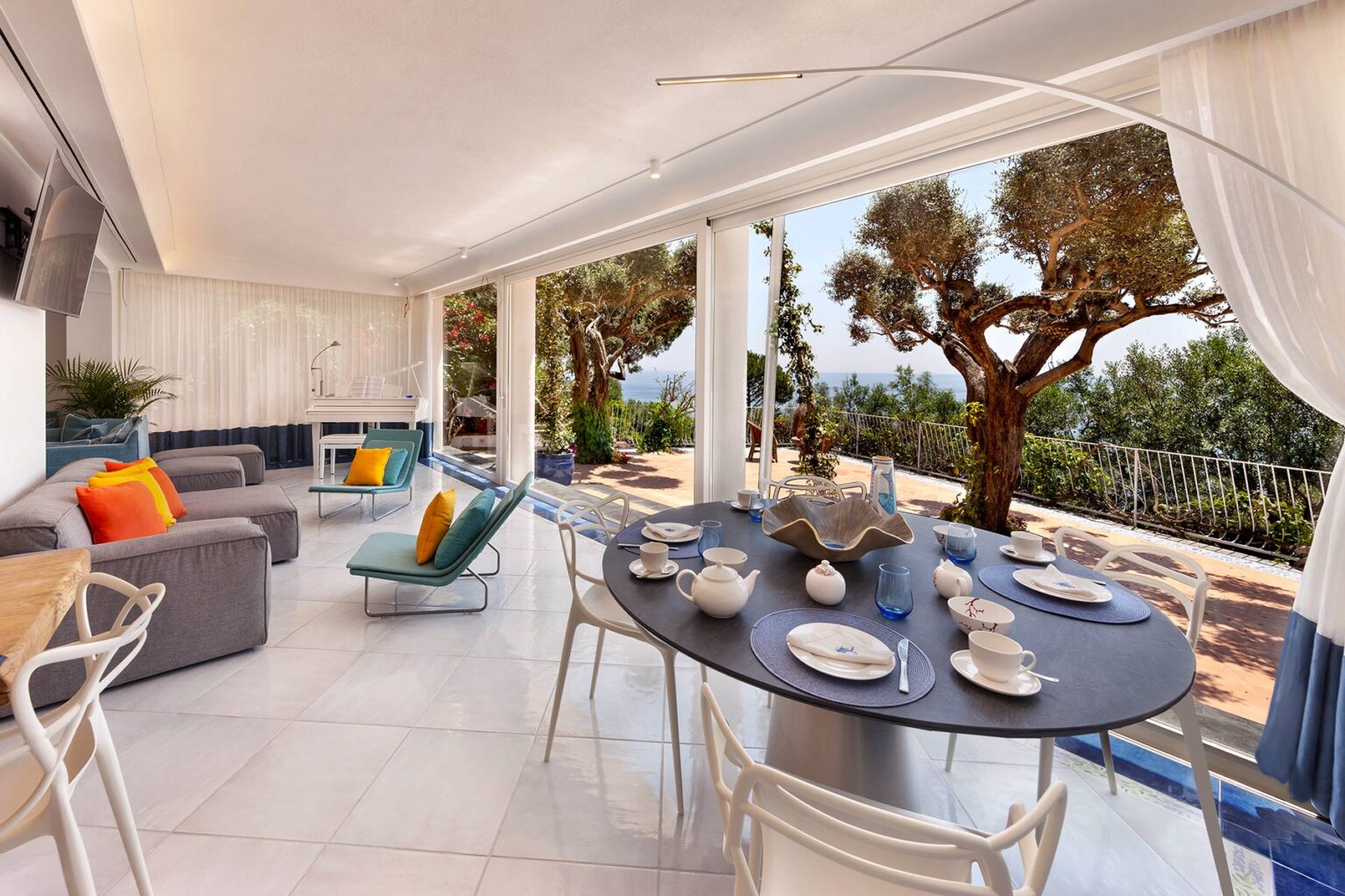 Modern dining area with a scenic view, featuring a round table set with dishes and cutlery, surrounded by stylish seating and large windows showcasing lush greenery outside. | Only on SopranoVillas