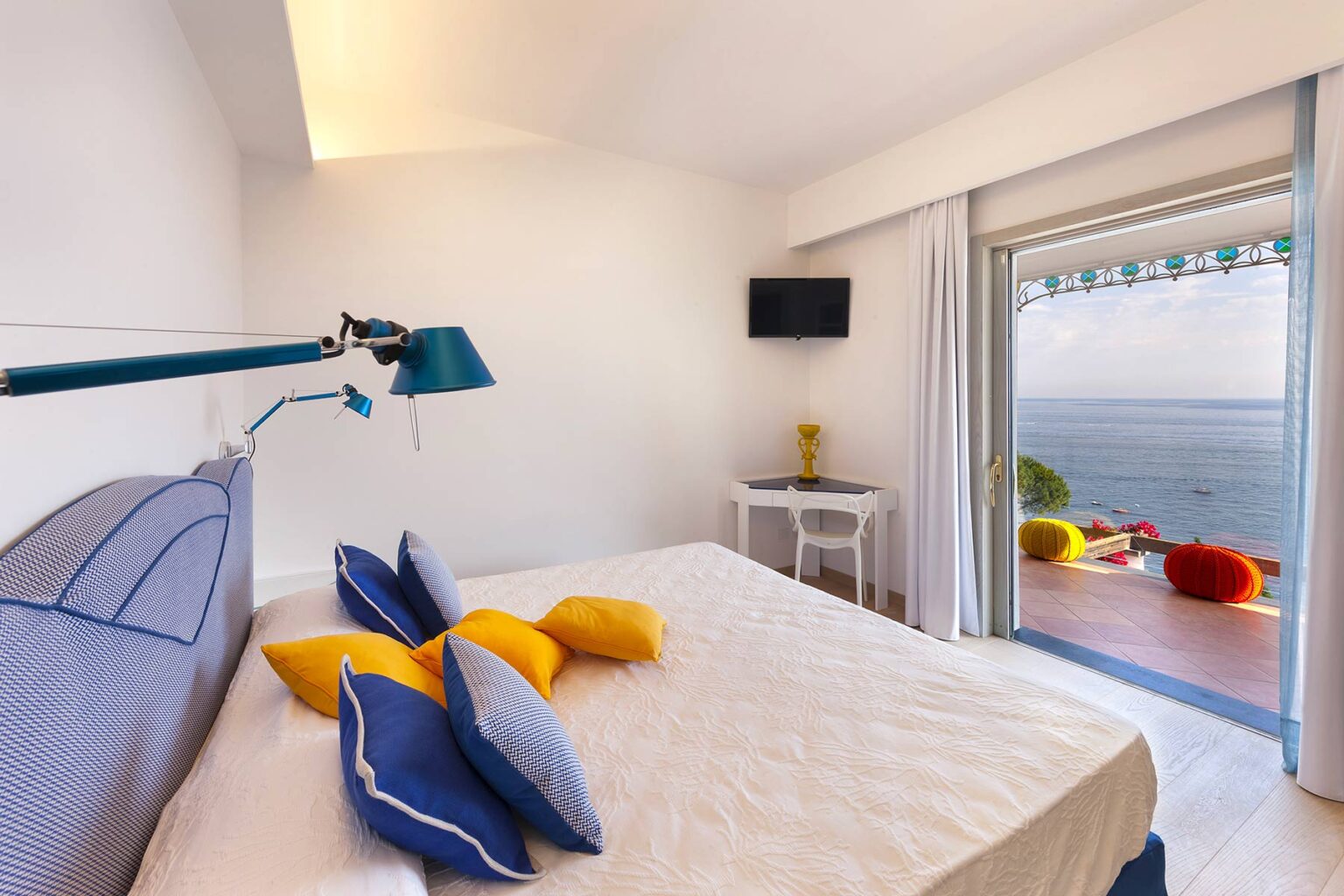 Modern hotel room with a cozy bed adorned with blue and yellow pillows, featuring a writing desk, a flat-screen TV, and a balcony overlooking the sea. Bright, airy design with natural light streaming in through large glass doors. | Only on SopranoVillas