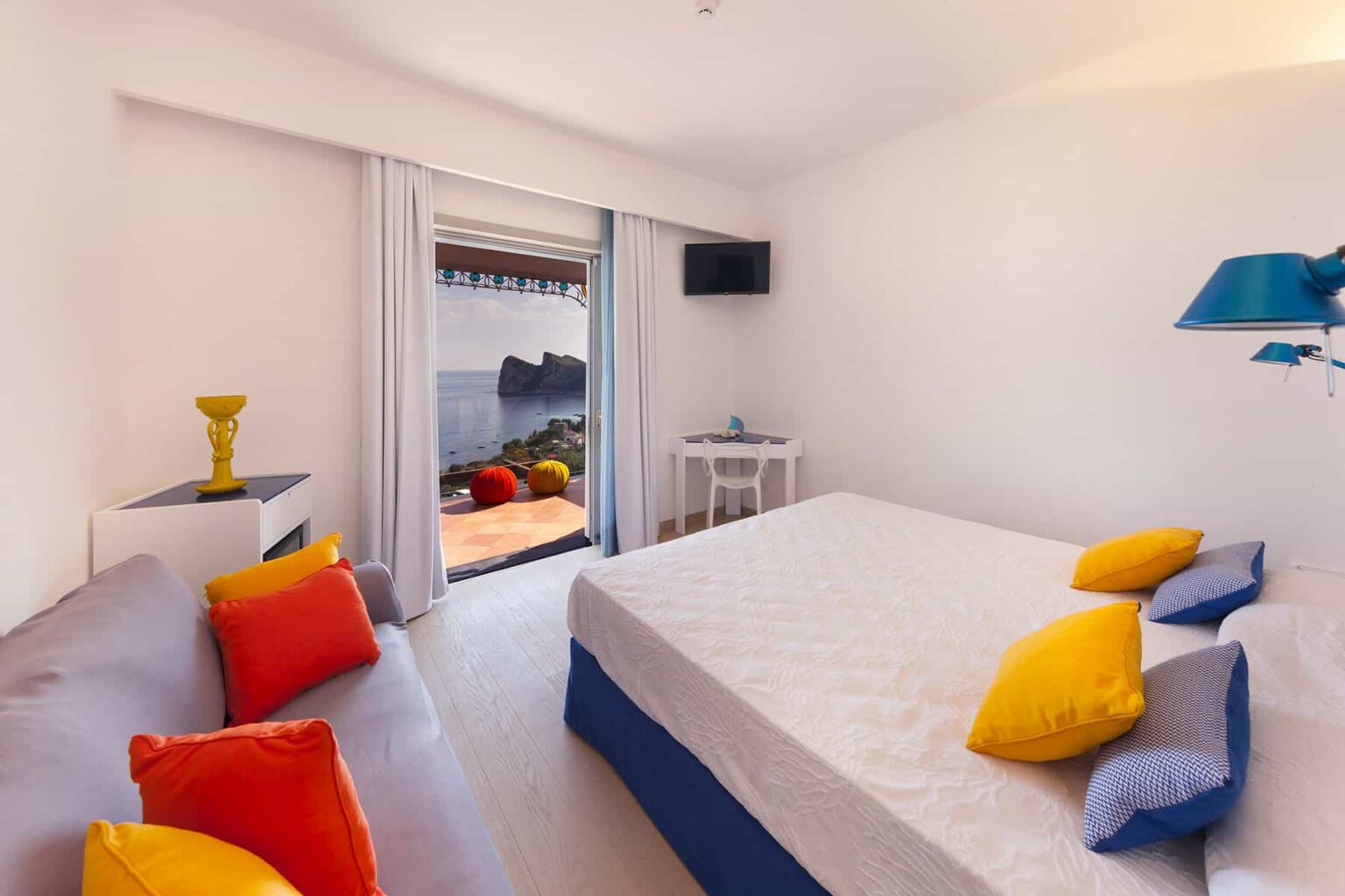 Bright and airy bedroom featuring a queen-sized bed with colorful cushions, a cozy sofa, and a small table. Large sliding doors open to a scenic outdoor area, enhancing the room's natural light and inviting ambiance. | Only on SopranoVillas