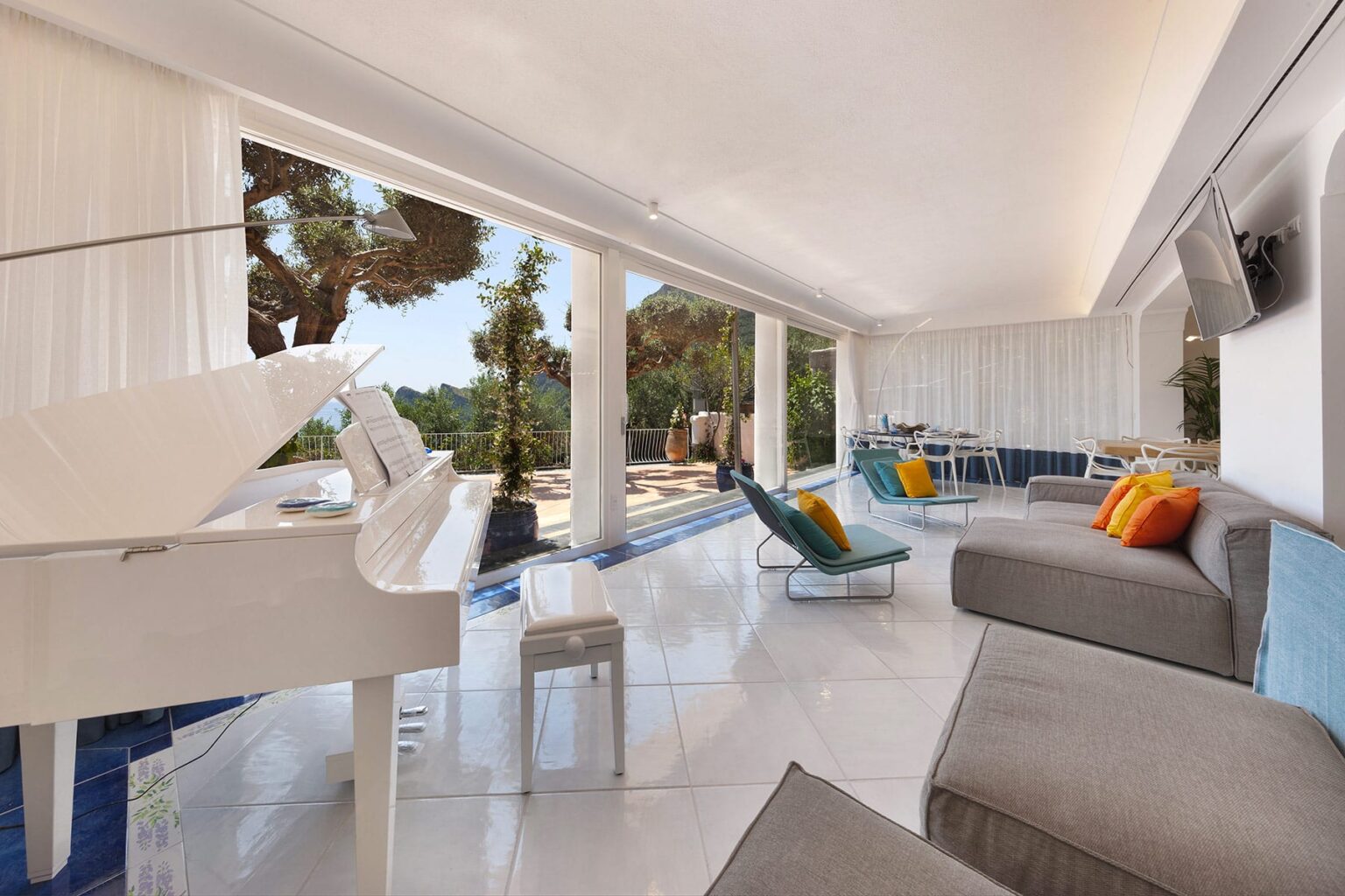 Bright and modern living room featuring a white piano, sleek furnishings, and colorful cushions, with large windows offering a view of the outdoor landscape and pool area. Ideal for luxury home design inspiration. | Only on SopranoVillas