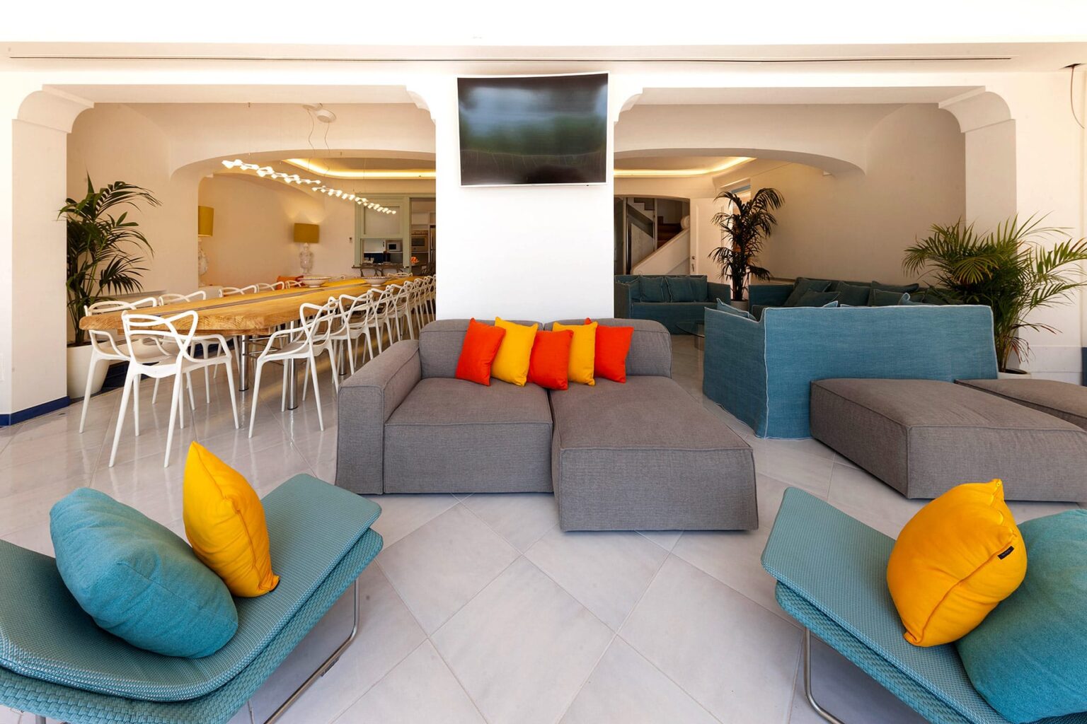 Modern lounge area featuring colorful cushions on gray sofas, stylish seating arrangements, and a dining space with white chairs. Bright interior design with plants and a flat-screen TV, creating a welcoming atmosphere. | Only on SopranoVillas