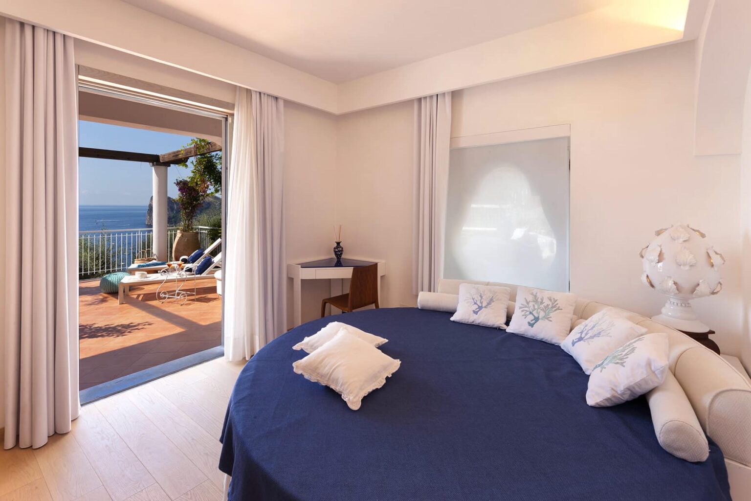 Modern bedroom with a round bed and blue bedding, featuring large windows that open to a terrace with ocean views. The room has light-colored walls, sheer curtains, and stylish decor, creating a serene and inviting atmosphere. | Only on SopranoVillas