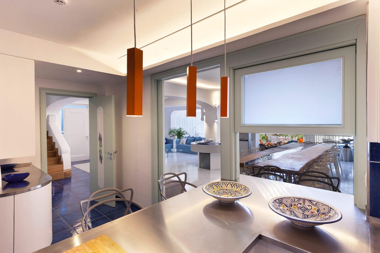 Modern kitchen interior featuring sleek stainless steel countertops, decorative bowls, and contemporary orange pendant lights. An open layout connects to a bright dining area with large windows, providing a view of the outdoor space. | Only on SopranoVillas