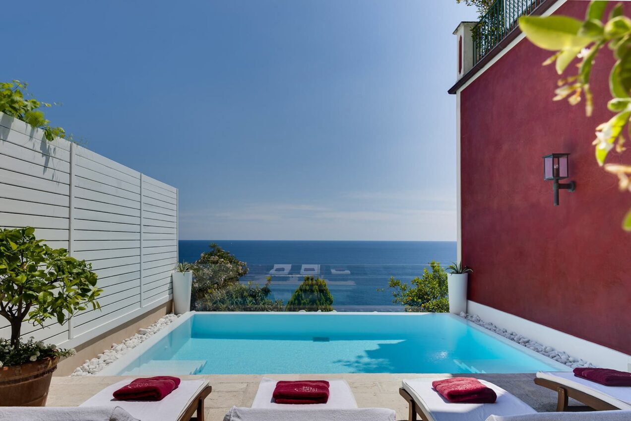 Luxurious outdoor pool with ocean view, surrounded by greenery and lounge chairs, perfect for relaxation and leisure. Bright blue sky enhances the stunning coastal scenery. | Only on SopranoVillas