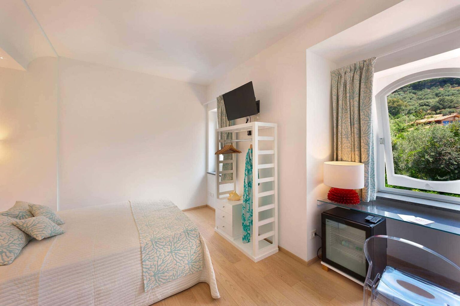 Cozy hotel room featuring a double bed, modern decor, a flat-screen TV, a mini-fridge, and a large window with scenic views. The space is well-lit with natural light and has stylish furnishings. | Only on SopranoVillas